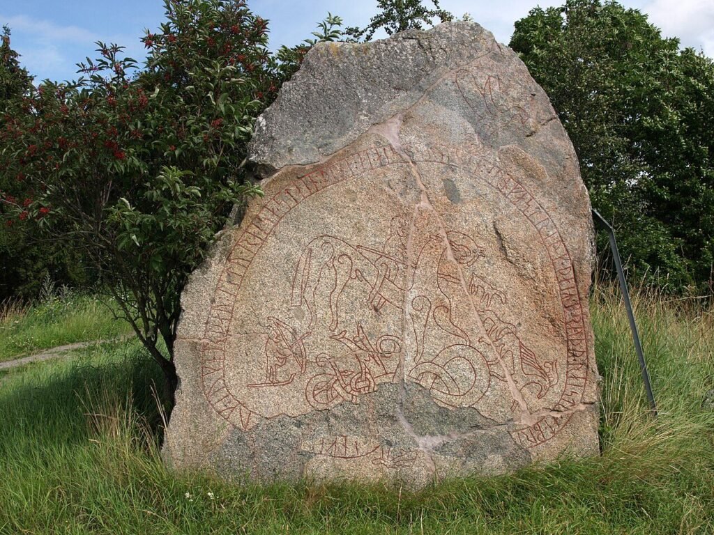 A large gray runestone carved with a band of runes, standing in a grassy field in Sweden